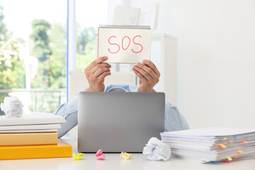 Man holding notebook with word SOS and hiding face behind laptop at table in office