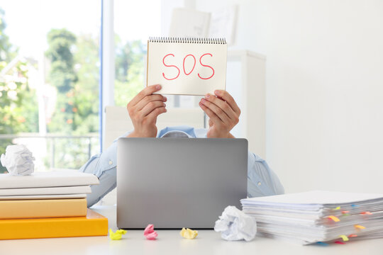 Man holding notebook with word SOS and hiding face behind laptop at table in office