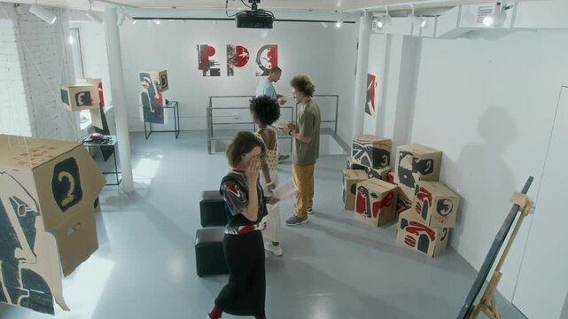 Wide high-angle shot of diverse people walking around conceptual art exhibition in museum, viewing paintings and installations on display, discussing artworks, checking artist information in brochures