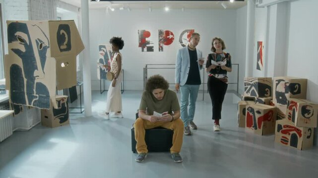 Wide timelapse shot of multiethnic visitors wandering around contemporary art exhibition in gallery - admiring artworks, using smartphone while sitting on pouffe, reading information leaflet