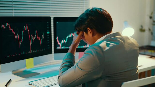 Young asian stock trader is feeling stressed while working at his desk. He is touching his head with his hand, seemingly having a loss on the stock market