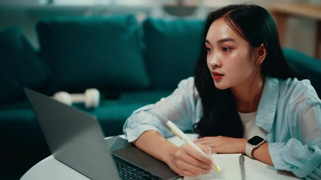 Young woman working from home, taking notes in a notebook while following an online course on her laptop. Focused and serious, she engages in remote learning and education