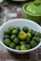 A bowl of small, green calamansi or songkit oranges, with one partially peeled to reveal the juicy yellow flesh. Citrusy and fresh.