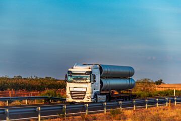 Truck transporting large steel pipes on a highway at sunrise, side view.