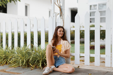 Relaxed young woman sitting against a white picket fence, enjoying her phone in a peaceful garden,...
