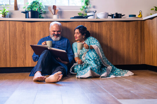 Joyful Indian couple relaxing with tea in a modern kitchen, looking through old memories - Powered by Adobe