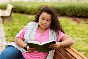 Obraz premium A young woman sits on a wooden bench in a park, absorbed in reading a book. She enjoys the warmth of the sun while surrounded by green grass and flowering plants.