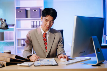 Asian Businessman analyzing reports in a modern office, working on a computer. Perfect for corporate, business, and over time concepts