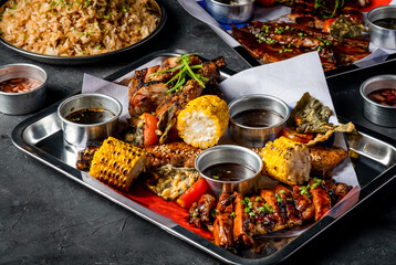 Filipino Feast with Grilled Meat, Garlic Fried Rice, grill corn cob, Peri Peri Chicken, Jerk Chicken, Beef Belly Brisket and Sauces served in tray on grey background closeup side view of platter