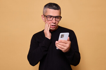 Shocked guy wearing glasses and jumper staring at smartphone feeling scared and sad while checking social media connected with technology standing indoor isolated over beige background