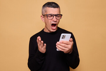 Angry man wearing glasses and jumper screaming while holding smartphone feeling aggressive about...