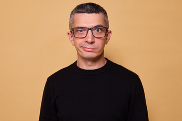 Funny man wearing glasses and black casual sweater standing indoors isolated over beige background with puzzled expression grimacing while pondering idea frowning face with foolish grimace