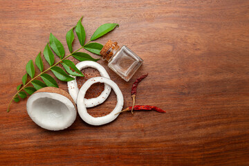 Top view of freshly cracked coconut fruit pieces, with Curry, leaves Red chili, and Coconut oil in a glass bottle, on a wooden background.