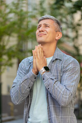 Caucasian mature man tourist praying with closed eyes to God asking for blessing, help, forgiveness outdoor. Grey-haired guy clasping hands wishing luck in urban city street. Town lifestyle. Vertical.