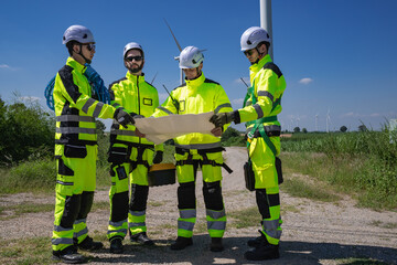 Maintenance engineer team standing at windmills at wind turbine farm. Group of people wear safety helmet and uniform working at alternative renewable energy wind station. Sustainable energy technology