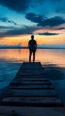 Fototapeta premium A man stands at the end of an endless pier above calm waters, gazing at the horizon where day meets night
