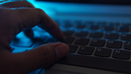someone's fingers typing on a laptop keyboard. dim background with blue light from the side....