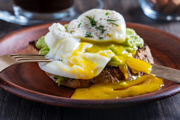 Cutting poached egg with runny egg yolk over bread toast with mashed avocado spread in plate, closeup. Healthy breakfast or lunch food