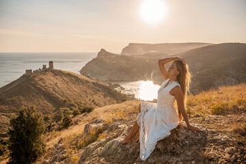 A woman in a white dress is sitting on a rock overlooking a body of water. She is enjoying the view and taking in the scenery.