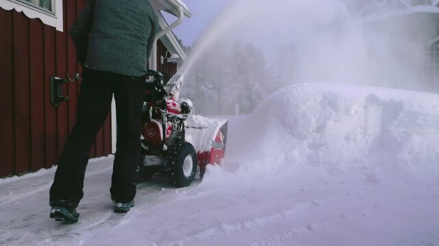 A man uses a snow blower to clear thick snow near a red wooden house during winter. Snow is being thrown high into the air as it clears the path in a snowy residential setting