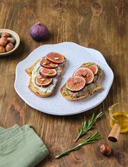 Two open sandwiches with fresh figs, ricotta and pate on a light ceramic plate on a wooden background. Recipes figs.
