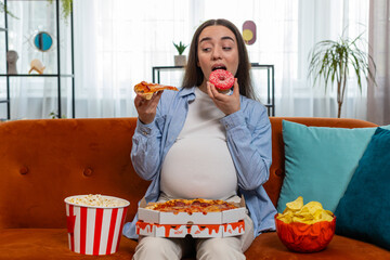 Pregnant woman eating unhealthy junk food pizza, popcorn crisp sitting on sofa in home living room....