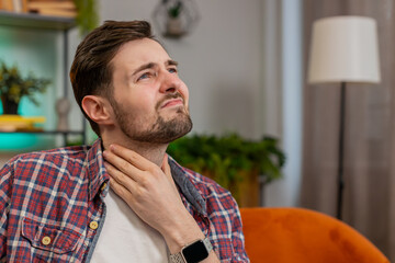 Naklejka premium Upset Caucasian man in plaid shirt holds hands over sore throat. Painful neck and frowning, thyroid disorders, suffering sore throat, tonsils inflammation. Young guy sitting on sofa at home room