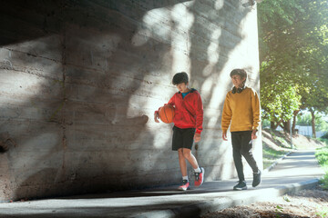 Two young boys are walking down a sidewalk, one of them holding a basketball
