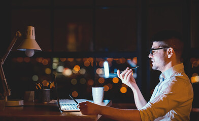 Business Lifestyle. Man Using Smartphone Voice Assistant Working On Laptop In Modern Office At Night. Copy Space