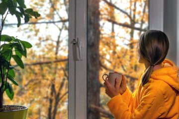 Woman in yellow warm robe by window in fall enjoying view of fall foliage in rain and hot drink