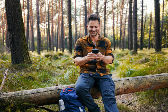 Happy man using mobile phone sitting in forest during weekend hike trip
