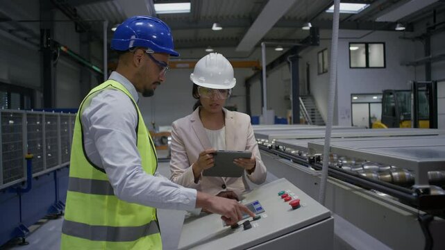 Female Indian factory supervisor is on-the-job training an African American male employee, in a blue helmet and yellow vest, to operate machinery.