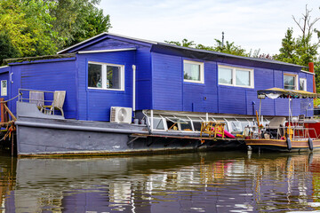 Houseboat with blue walls anchored on bank of shipping canal outside the city of Maastricht, green foliage of trees in background, cloudy day in South Limburg, Netherlands