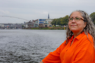 Fototapeta premium Adult woman with dejected expression sailing on tourist boat on Maas river, modern bridge and buildings on blurred background, cloudy day in Maastricht city in South Limburg, Netherlands