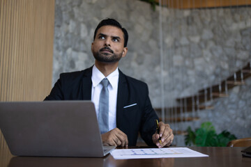 A handsome businessman in a suit sitting at a desk, working intently on his laptop, showcasing professionalism and success in a modern office environment