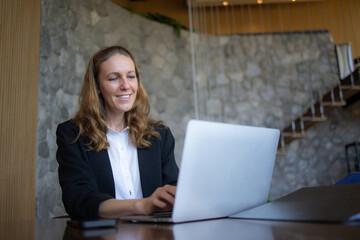 Confident businesswoman in a suit smiling while working on a laptop and document file  at her desk in an office setting