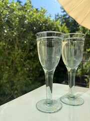 Two glasses filled with cool drinks standing on a white table in a garden or a terrace. Sun is shining. In the background, there is a green hedge and a parasol.