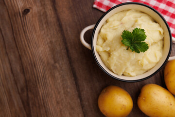 Mashed potatoes in bowl surrounded by fresh potatoes and red napkin, brown wooden background, top view