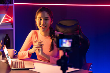 Smiling creative young beautiful Asian influencer recording on camera with positive podcast speech at night time, using laptop for presentation online at pink neon light modern studio. Stratagem.
