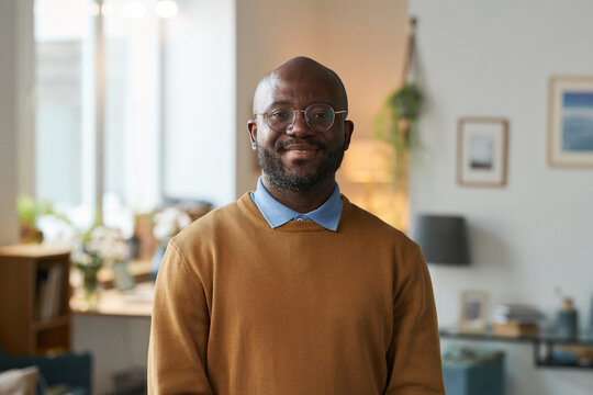 Front view portrait of adult African American man wearing glasses and smiling a camera standing in warm home setting copy space