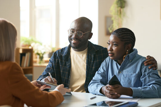 Portrait of smiling African American couple enjoying consultation with insurance agent sitting at desk in office and signing forms copy space
