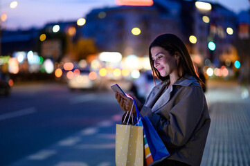 Elegant woman browsing on smartphone after shopping spree in city at night