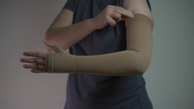 Prophylactics of thrombosis. Woman removes compression sleeve from left hand on gray background. Effective anti-thrombosis therapy involves compression sleeves to improve vein health and blood flow. 