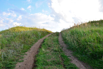 a dirt country road leads to a grassy hill with the light in the background