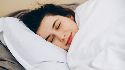 Cozy bedtime. Good morning. Healthy sleep. Calm smiling woman closed eyes under blanket soft white bed sheet enjoying bedroom comfort.