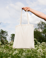 Mature woman, female hand holding white bag canvas fabric for mock up blank template on meadow with flowers background. Ecology, reusable concept. Eco friendly tote shopper