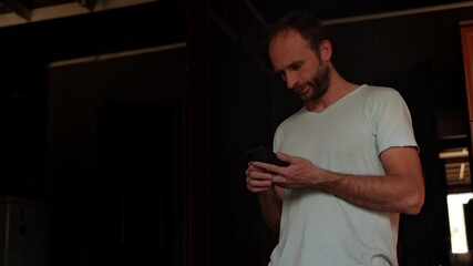 A bearded man sits at home in the morning, engrossed in his mobile phone as he enthusiastically reads the news. His expressions convey interest and excitement about what he's discovering
