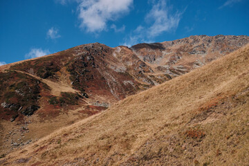 A breathtaking view of a mountain range with a blue sky and white clouds. The mountain peaks are covered in grass and rocks. Făgăraș Mountains, Romania