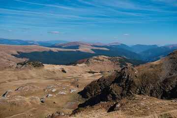 Panoramic view of a mountain range with a blue sky. The mountains are covered in grass and rocks. Parâng Mountains, Romania