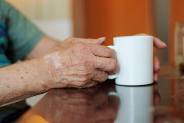 An elderly person hands are gently resting on a table near a white mug.